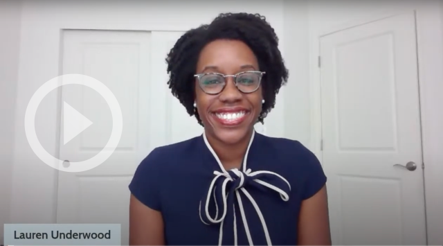 Congresswoman Lauren Underwood smiles at the camera. A transparent "play" button is on the left.